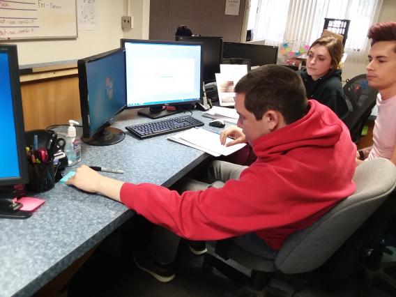 male student reaching for post-it note on desk next to computer