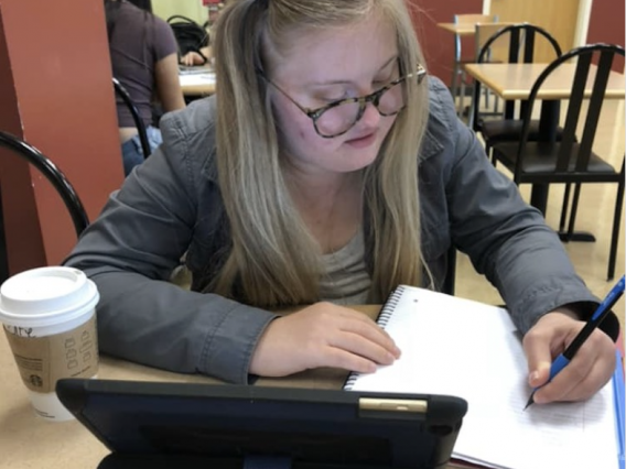 female student writing in notebook while using tablet propped up on table